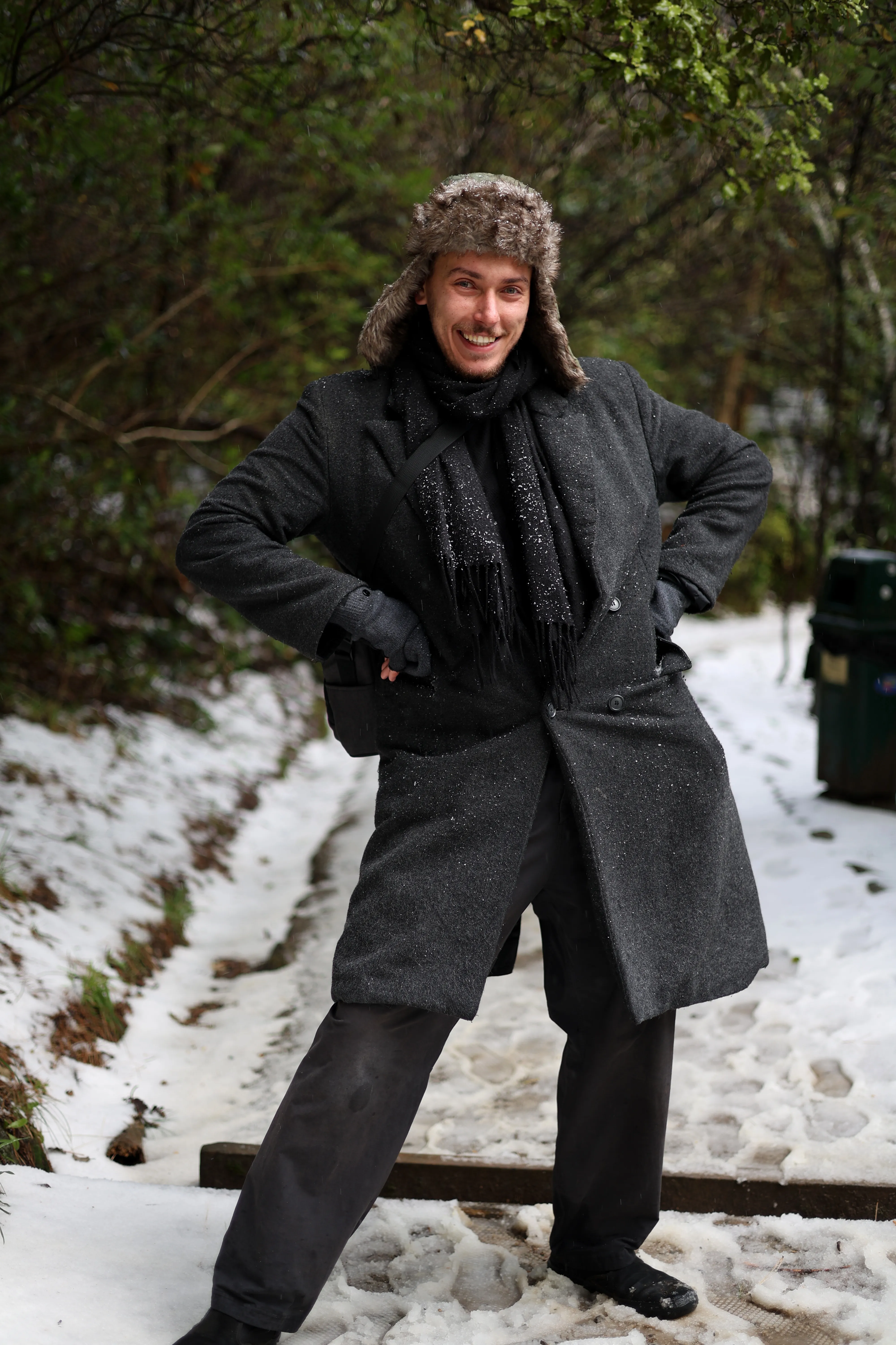 Kenan-John smiling with his hands on his hips, wearing a dark coat and fur hat on a snowy path